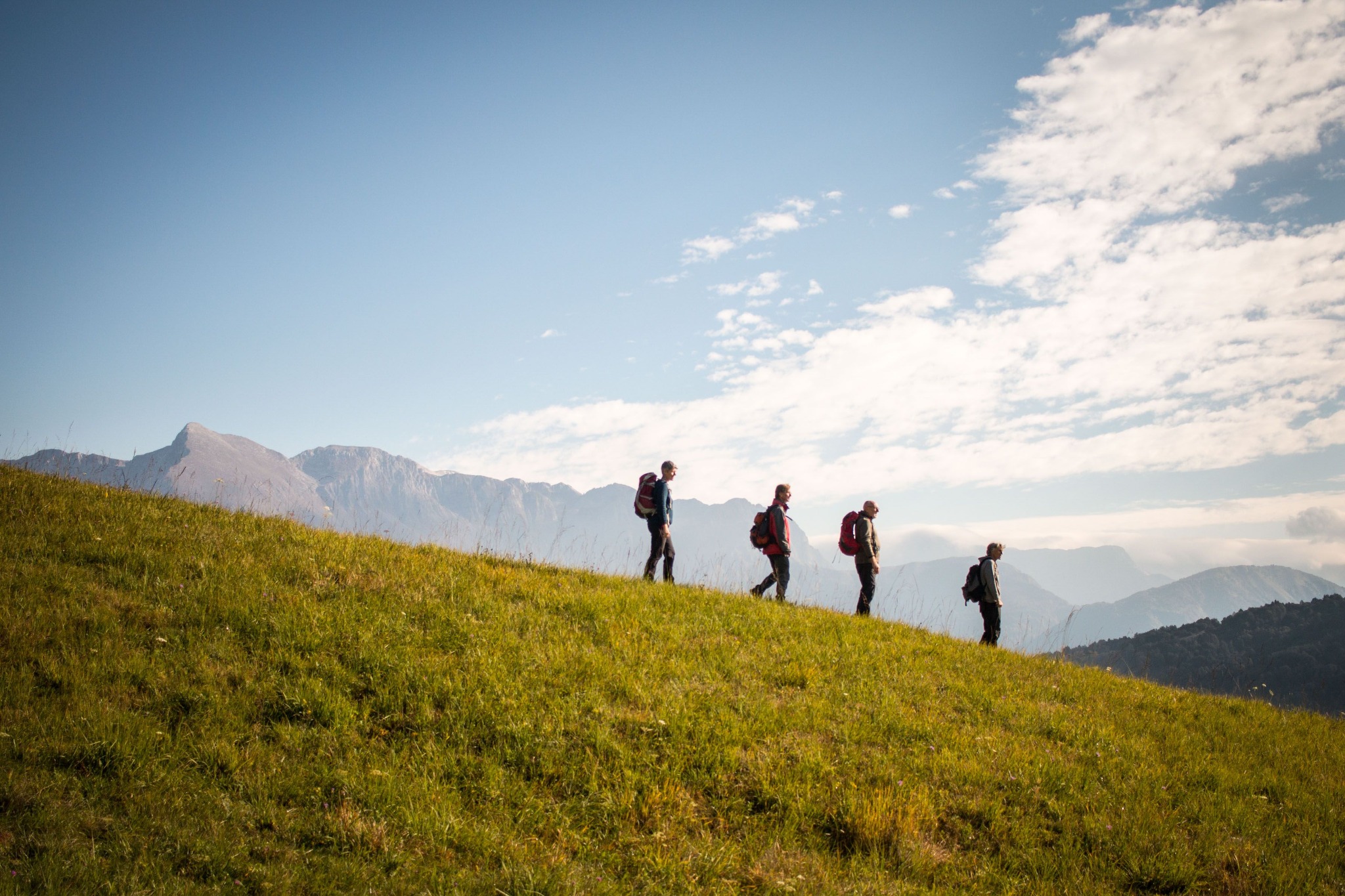 Four walkers with rucksacks crossing a grassy hillside, with mountains in the background.